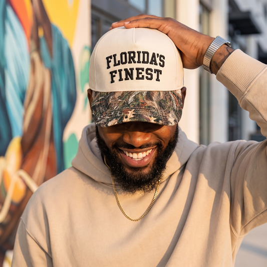Smiling man wearing a cream hoodie and a two-tone Florida’s Finest hat with a camouflage brim, posing outdoors in warm sunlight.