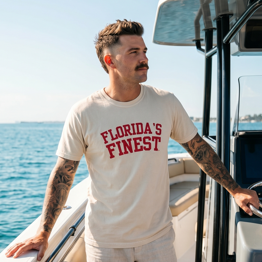 Man wearing a Florida’s Finest graphic t-shirt while standing on a boat in open water. Florida lifestyle apparel featuring bold red collegiate-style lettering, perfect for boating, fishing, beach days, and coastal living.