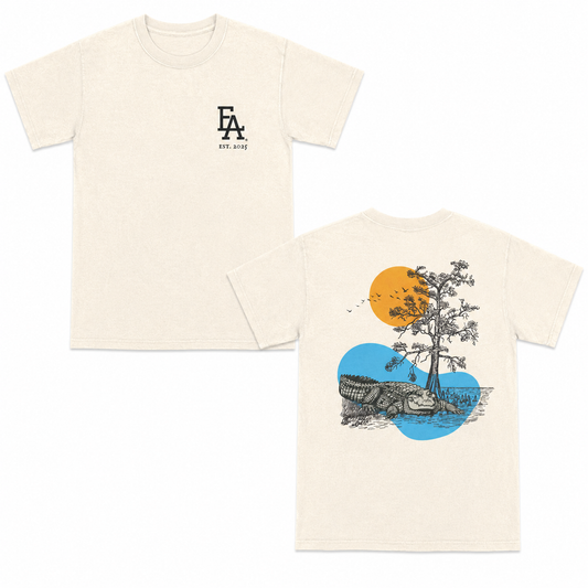 Ivory short-sleeve T-shirt with a minimalist black “FLA est. 2025” logo on the front and a detailed back graphic featuring a Florida alligator, tree, birds, and sun over water in a vintage outdoor illustration style.