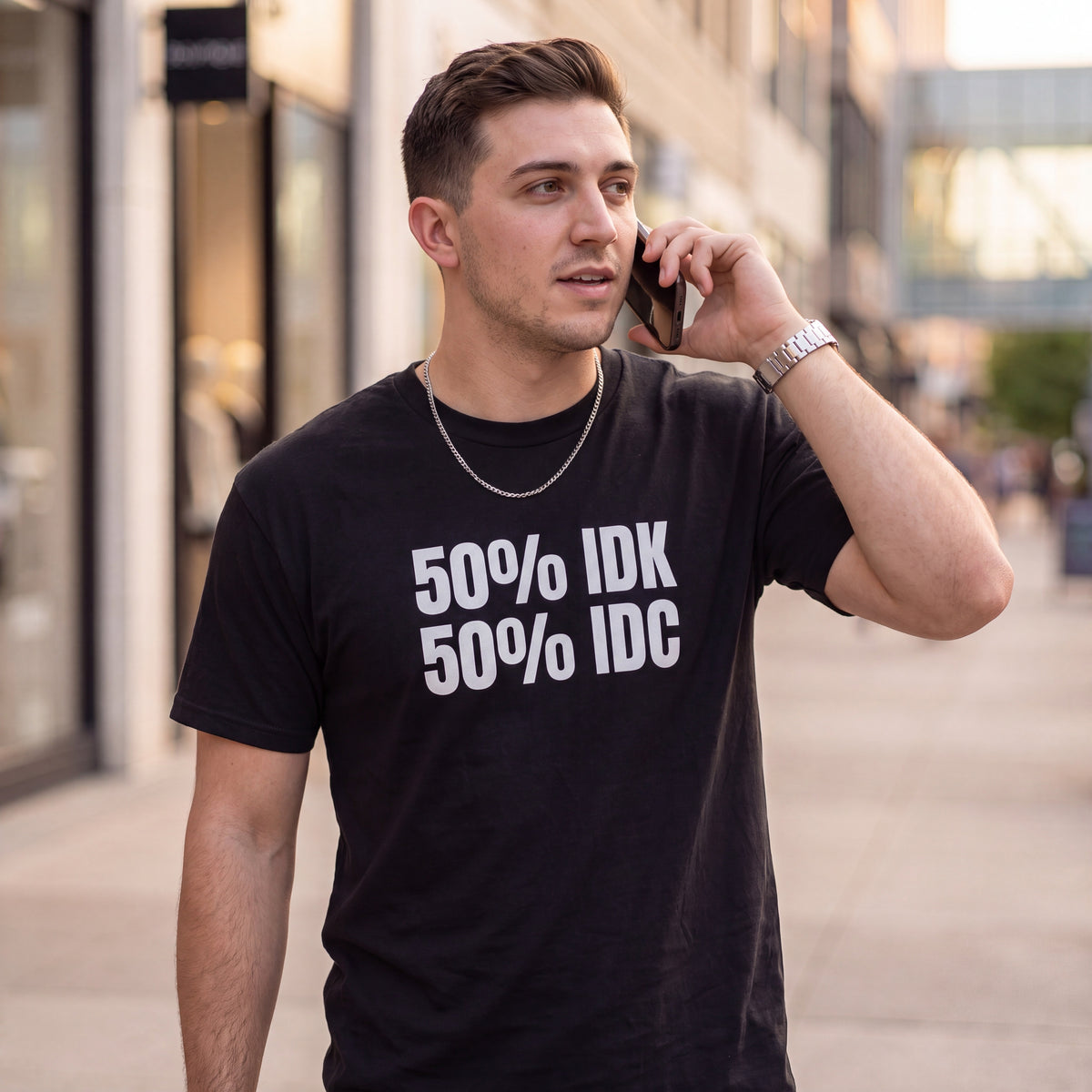 Man walking outdoors while talking on a phone, wearing a black graphic t-shirt with bold white text reading “50% IDK 50% IDC.” He wears a silver chain and watch in a modern urban shopping district, creating a casual lifestyle look for the humorous statement tee.