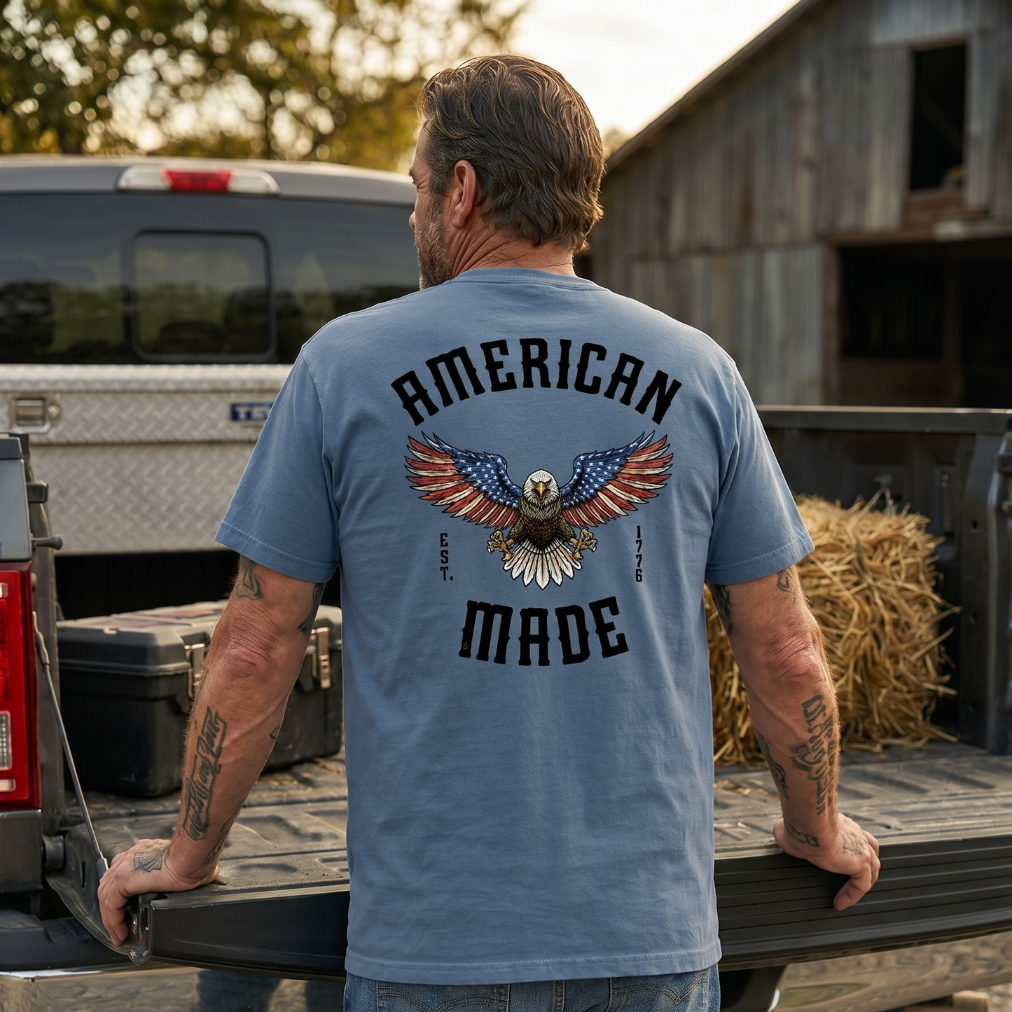 Man standing at the tailgate of a pickup truck wearing a blue patriotic t-shirt featuring a distressed bald eagle with American flag wings and the text “American Made” with “Est. 1776.” Rustic farm setting highlights American-made apparel, patriotism, freedom, hardworking values, rural lifestyle, and pride in the USA.