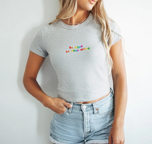 Woman wearing a gray baby tee with colorful text and light blue jeans against a white background