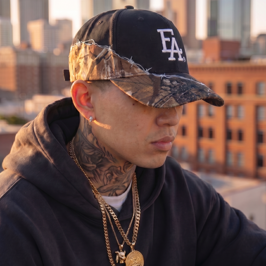 Model wearing a black and camo barbed wire baseball cap featuring a bold white embroidered FLA® logo. Structured curved-brim hat with camouflage visor, barbed wire detail, and urban streetwear styling against a city skyline backdrop.