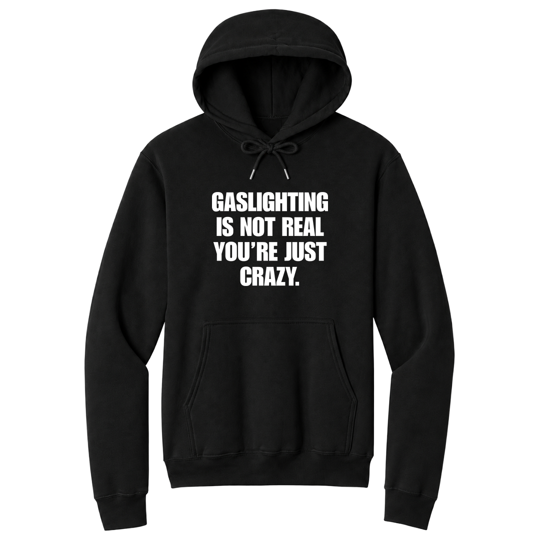 Front view of a black pullover hoodie featuring the phrase "GASLIGHTING IS NOT REAL YOU’RE JUST CRAZY." printed in bold white uppercase lettering centered on the chest. The hoodie includes a front kangaroo pocket and matching black drawstrings tied in a bow, with the minimalist black-and-white design creating a bold, sarcastic statement look.