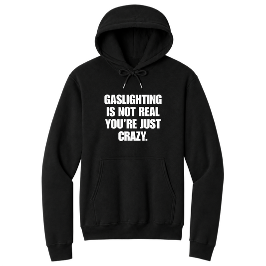 Front view of a black pullover hoodie featuring the phrase "GASLIGHTING IS NOT REAL YOU’RE JUST CRAZY." printed in bold white uppercase lettering centered on the chest. The hoodie includes a front kangaroo pocket and matching black drawstrings tied in a bow, with the minimalist black-and-white design creating a bold, sarcastic statement look.