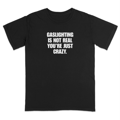 Front view of a black short sleeve t-shirt featuring the phrase "GASLIGHTING IS NOT REAL YOU’RE JUST CRAZY." printed in bold white uppercase lettering centered on the chest. The high-contrast black-and-white design creates a clean, minimalist statement tee with a humorous, sarcastic style.