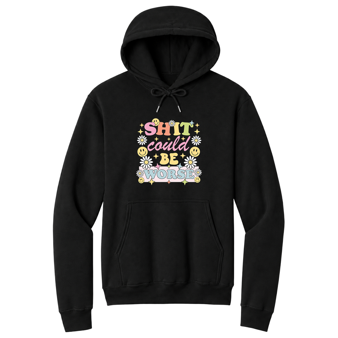 Front view of a black pullover hoodie featuring a colorful retro-inspired graphic with the phrase "SHIT could BE WORSE" in playful pastel typography. The design is surrounded by white daisies, yellow smiley faces, and sparkling star accents, creating a cheerful vintage aesthetic that humorously contrasts with the sarcastic message. The drawstrings are tied into a short bow above the design for a realistic product mockup. A funny adult hoodie with bold humor, retro floral graphics, and casual style