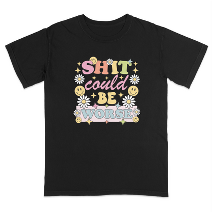Front view of a black graphic t-shirt featuring a retro-inspired pastel design with the phrase "SHIT could BE WORSE" in colorful vintage lettering. The design is decorated with white daisies, yellow smiley faces, and sparkling star accents, creating a playful 70s aesthetic that contrasts with the humorous sarcastic message. A funny adult graphic tee with retro floral artwork, pastel colors, and bold statement typography for casual everyday wear.
