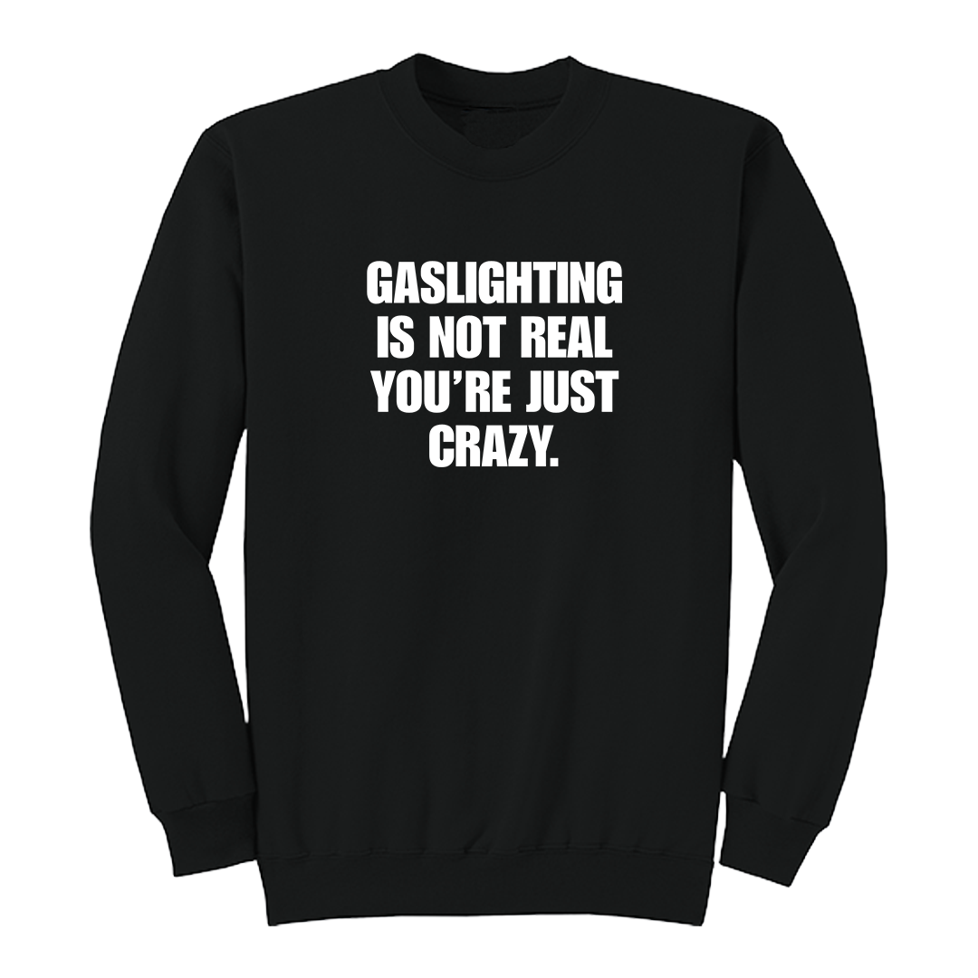 Front view of a black crewneck sweatshirt featuring the phrase "GASLIGHTING IS NOT REAL YOU’RE JUST CRAZY." printed in bold white uppercase lettering centered on the chest. The clean black-and-white design creates a minimalist statement sweatshirt with a bold, sarcastic aesthetic.