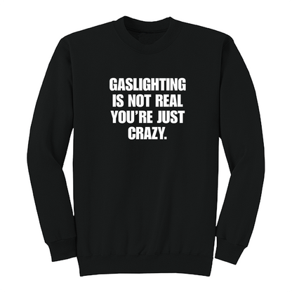 Front view of a black crewneck sweatshirt featuring the phrase "GASLIGHTING IS NOT REAL YOU’RE JUST CRAZY." printed in bold white uppercase lettering centered on the chest. The clean black-and-white design creates a minimalist statement sweatshirt with a bold, sarcastic aesthetic.