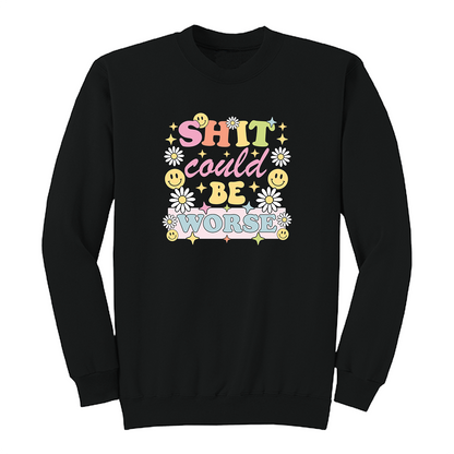 Front view of a black long sleeve graphic shirt featuring a colorful retro-inspired design with the phrase "SHIT could BE WORSE" in playful pastel typography. The graphic is surrounded by white daisies, yellow smiley faces, and sparkling star accents, creating a cheerful vintage aesthetic that humorously contrasts with the sarcastic message. A funny adult long sleeve shirt with retro floral graphics, bold humor, and casual streetwear style.