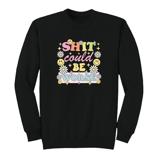 Front view of a black long sleeve graphic shirt featuring a colorful retro-inspired design with the phrase "SHIT could BE WORSE" in playful pastel typography. The graphic is surrounded by white daisies, yellow smiley faces, and sparkling star accents, creating a cheerful vintage aesthetic that humorously contrasts with the sarcastic message. A funny adult long sleeve shirt with retro floral graphics, bold humor, and casual streetwear style.