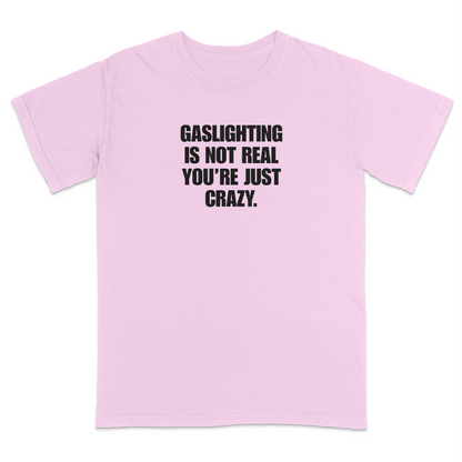 Front view of a light pink short sleeve t-shirt featuring the phrase "GASLIGHTING IS NOT REAL YOU’RE JUST CRAZY." printed in bold black uppercase lettering centered on the chest. The high-contrast black-and-white design creates a clean, minimalist statement tee with a humorous, sarcastic style.