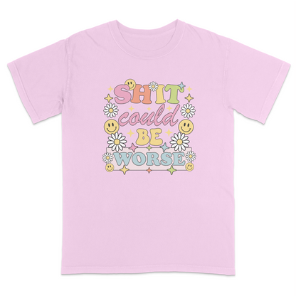 Front view of a pink graphic t-shirt featuring a retro-inspired pastel design with the phrase "SHIT could BE WORSE" in colorful vintage lettering. The design is decorated with white daisies, yellow smiley faces, and sparkling star accents, creating a playful 70s aesthetic that contrasts with the humorous sarcastic message. A funny adult graphic tee with retro floral artwork, pastel colors, and bold statement typography for casual everyday wear.