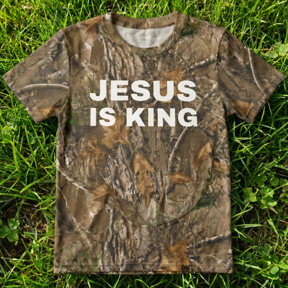 Camo performance t-shirt featuring bold white "Jesus Is King" text on a realistic woodland camouflage pattern. Faith-based Christian apparel designed for hunting, outdoor activities, church events, and everyday wear.