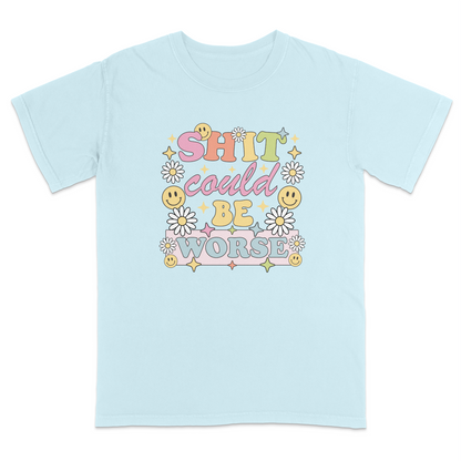 Front view of a chambray graphic t-shirt featuring a retro-inspired pastel design with the phrase "SHIT could BE WORSE" in colorful vintage lettering. The design is decorated with white daisies, yellow smiley faces, and sparkling star accents, creating a playful 70s aesthetic that contrasts with the humorous sarcastic message. A funny adult graphic tee with retro floral artwork, pastel colors, and bold statement typography for casual everyday wear.