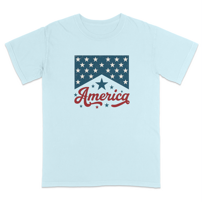 Light blue patriotic graphic t-shirt featuring a vintage-inspired red “America” script with blue stars and retro Americana design for classic Fourth of July and USA-themed style.