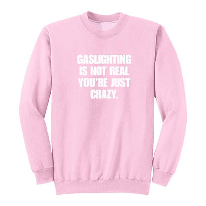Front view of a light pink crewneck sweatshirt featuring the phrase "GASLIGHTING IS NOT REAL YOU’RE JUST CRAZY." printed in bold white uppercase lettering centered on the chest. The clean black-and-white design creates a minimalist statement sweatshirt with a bold, sarcastic aesthetic.