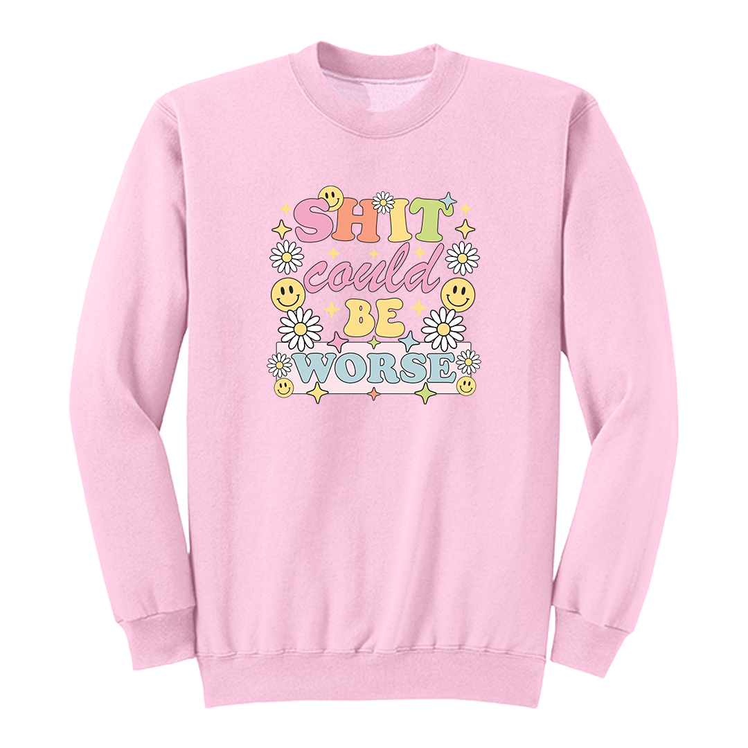 Front view of a pink long sleeve graphic shirt featuring a colorful retro-inspired design with the phrase "SHIT could BE WORSE" in playful pastel typography. The graphic is surrounded by white daisies, yellow smiley faces, and sparkling star accents, creating a cheerful vintage aesthetic that humorously contrasts with the sarcastic message. A funny adult long sleeve shirt with retro floral graphics, bold humor, and casual streetwear style.