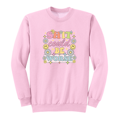 Front view of a pink long sleeve graphic shirt featuring a colorful retro-inspired design with the phrase "SHIT could BE WORSE" in playful pastel typography. The graphic is surrounded by white daisies, yellow smiley faces, and sparkling star accents, creating a cheerful vintage aesthetic that humorously contrasts with the sarcastic message. A funny adult long sleeve shirt with retro floral graphics, bold humor, and casual streetwear style.