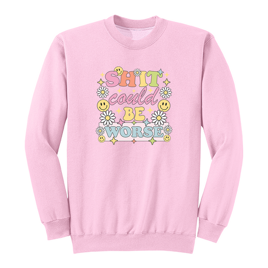 Front view of a pink long sleeve graphic shirt featuring a colorful retro-inspired design with the phrase "SHIT could BE WORSE" in playful pastel typography. The graphic is surrounded by white daisies, yellow smiley faces, and sparkling star accents, creating a cheerful vintage aesthetic that humorously contrasts with the sarcastic message. A funny adult long sleeve shirt with retro floral graphics, bold humor, and casual streetwear style.