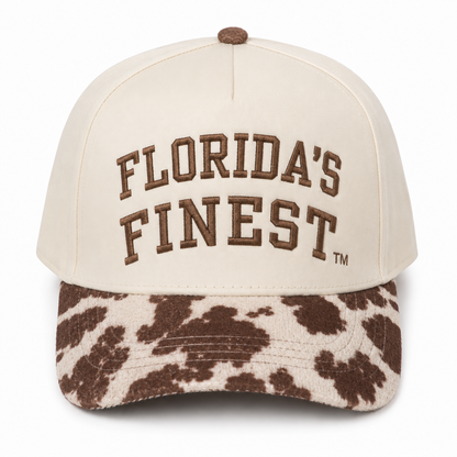 Front view of a cream snapback hat featuring raised brown "Florida's Finest" embroidery, paired with a brown and cream cow print curved bill. The structured cap is shown on a white background, highlighting the western-inspired design and premium embroidered details.