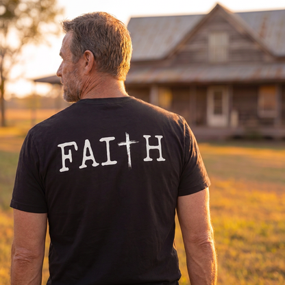 Man wearing a black Christian faith t-shirt featuring a large "FAITH" graphic with a cross design on the back, standing outdoors near a rustic farmhouse at sunset. Inspirational faith-based apparel for church, worship, and everyday Christian living.