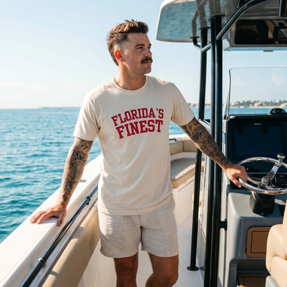 Man wearing a Florida’s Finest graphic t-shirt while standing on a boat in open water. Florida lifestyle apparel featuring bold red collegiate-style lettering, perfect for boating, fishing, beach days, and coastal living.
