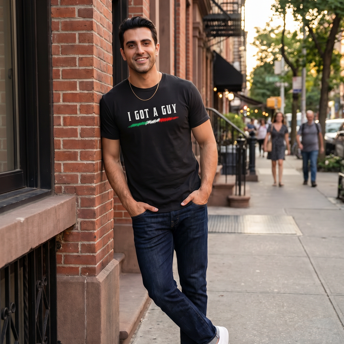 Man wearing a black “I Got A Guy” graphic t-shirt with an Italian flag accent, leaning against a brick building on a city street. Funny Italian heritage shirt celebrating Italian culture, connections, family pride, and casual everyday style.