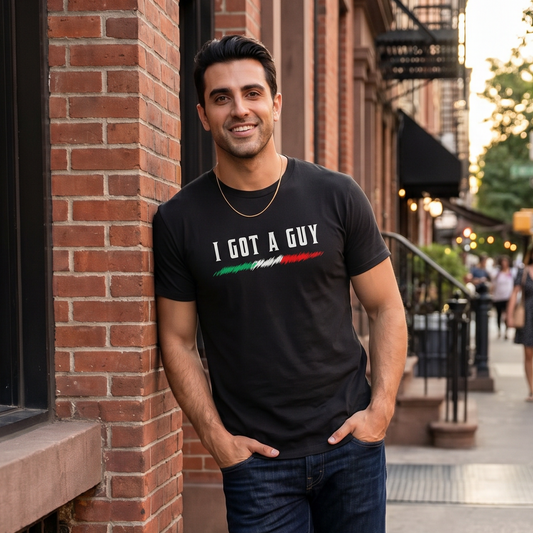 Man wearing a black “I Got A Guy” graphic t-shirt with an Italian flag accent, leaning against a brick building on a city street. Funny Italian heritage shirt celebrating Italian culture, connections, family pride, and casual everyday style.