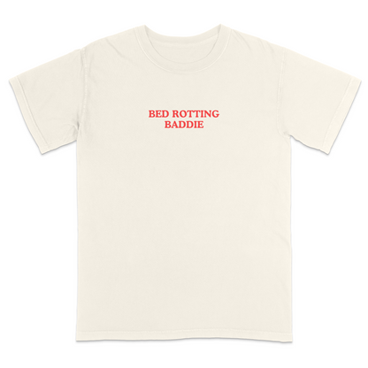 Front view of a ivory graphic T-shirt featuring the phrase "BED ROTTING BADDIE" in small vintage-style red lettering centered on the chest. This minimalist, trendy tee combines clean aesthetics with humorous Gen Z-inspired text for a casual everyday look.
