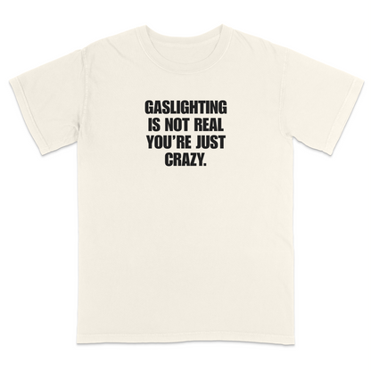 Front view of a ivory short sleeve t-shirt featuring the phrase "GASLIGHTING IS NOT REAL YOU’RE JUST CRAZY." printed in bold black uppercase lettering centered on the chest. The high-contrast black-and-white design creates a clean, minimalist statement tee with a humorous, sarcastic style.