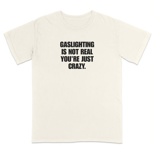 Front view of a ivory short sleeve t-shirt featuring the phrase "GASLIGHTING IS NOT REAL YOU’RE JUST CRAZY." printed in bold black uppercase lettering centered on the chest. The high-contrast black-and-white design creates a clean, minimalist statement tee with a humorous, sarcastic style.