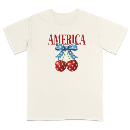 Ivory patriotic graphic t-shirt featuring red “AMERICA” text with star-pattern cherries and a blue bow design, combining vintage Americana style with playful patriotic fashion.