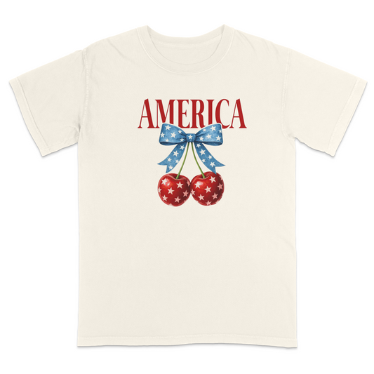 Ivory patriotic graphic t-shirt featuring red “AMERICA” text with star-pattern cherries and a blue bow design, combining vintage Americana style with playful patriotic fashion.