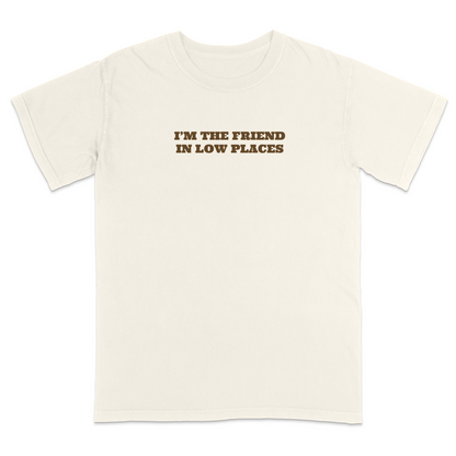 Front view of an ivory graphic T-shirt featuring the phrase "I'M THE FRIEND IN LOW PLACES" in bold vintage-style brown lettering centered across the chest. This western-inspired graphic tee has a clean minimalist design with a country music vibe, making it perfect for concerts, tailgates, casual outings, and everyday wear.
