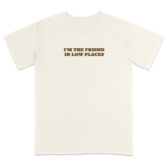 Front view of an ivory graphic T-shirt featuring the phrase "I'M THE FRIEND IN LOW PLACES" in bold vintage-style brown lettering centered across the chest. This western-inspired graphic tee has a clean minimalist design with a country music vibe, making it perfect for concerts, tailgates, casual outings, and everyday wear.