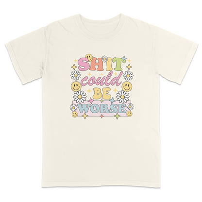Front view of a ivory graphic t-shirt featuring a retro-inspired pastel design with the phrase "SHIT could BE WORSE" in colorful vintage lettering. The design is decorated with white daisies, yellow smiley faces, and sparkling star accents, creating a playful 70s aesthetic that contrasts with the humorous sarcastic message. A funny adult graphic tee with retro floral artwork, pastel colors, and bold statement typography for casual everyday wear.