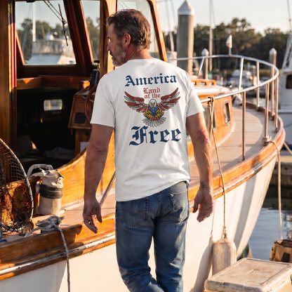 Man walking along a dock beside a classic wooden boat wearing a white patriotic t-shirt featuring a vintage eagle graphic and the phrase “America – Land of the Free.” Coastal marina setting highlights American pride, freedom, patriotic apparel, boating lifestyle, nautical adventure, and classic Americana style.