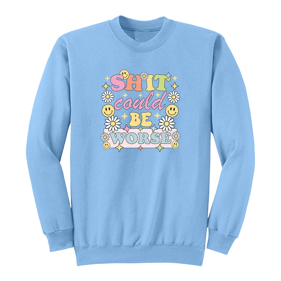 Front view of a light blue long sleeve graphic shirt featuring a colorful retro-inspired design with the phrase "SHIT could BE WORSE" in playful pastel typography. The graphic is surrounded by white daisies, yellow smiley faces, and sparkling star accents, creating a cheerful vintage aesthetic that humorously contrasts with the sarcastic message. A funny adult long sleeve shirt with retro floral graphics, bold humor, and casual streetwear style.