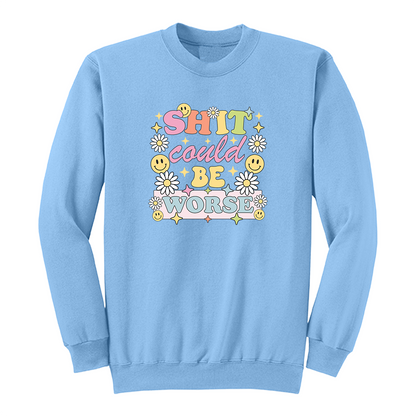 Front view of a light blue long sleeve graphic shirt featuring a colorful retro-inspired design with the phrase "SHIT could BE WORSE" in playful pastel typography. The graphic is surrounded by white daisies, yellow smiley faces, and sparkling star accents, creating a cheerful vintage aesthetic that humorously contrasts with the sarcastic message. A funny adult long sleeve shirt with retro floral graphics, bold humor, and casual streetwear style.