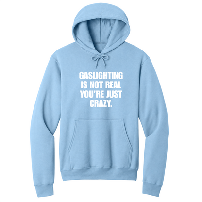 Front view of a light blue pullover hoodie featuring the phrase "GASLIGHTING IS NOT REAL YOU’RE JUST CRAZY." printed in bold white uppercase lettering centered on the chest. The hoodie includes a front kangaroo pocket and matching black drawstrings tied in a bow, with the minimalist black-and-white design creating a bold, sarcastic statement look.