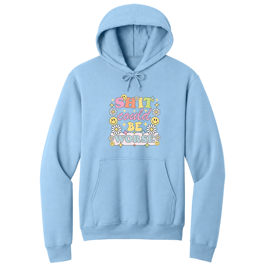 Front view of a light blue pullover hoodie featuring a colorful retro-inspired graphic with the phrase "SHIT could BE WORSE" in playful pastel typography. The design is surrounded by white daisies, yellow smiley faces, and sparkling star accents, creating a cheerful vintage aesthetic that humorously contrasts with the sarcastic message. The drawstrings are tied into a short bow above the design for a realistic product mockup. A funny adult hoodie with bold humor, retro floral graphics, and casual style