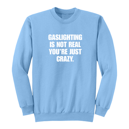 Front view of a light blue crewneck sweatshirt featuring the phrase "GASLIGHTING IS NOT REAL YOU’RE JUST CRAZY." printed in bold white uppercase lettering centered on the chest. The clean black-and-white design creates a minimalist statement sweatshirt with a bold, sarcastic aesthetic.