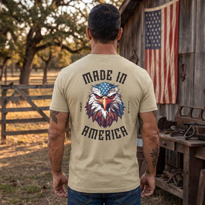 Man standing outdoors in a rustic farm setting wearing a tan patriotic t-shirt featuring a bold bald eagle graphic with American flag accents and the text “Made In America” with “Est. 1776.” Barn and American flag backdrop emphasize American pride, freedom, rural heritage, patriotic apparel, USA-made values, and country lifestyle.