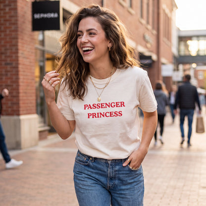 Lifestyle photo of a smiling woman walking through an outdoor shopping district wearing an ivory graphic T-shirt with the words "PASSENGER PRINCESS" printed in bold red vintage-style lettering across the chest. She pairs the casual tee with blue jeans and layered gold jewelry, creating a relaxed everyday outfit perfect for road trips, errands, vacations, and casual outings.