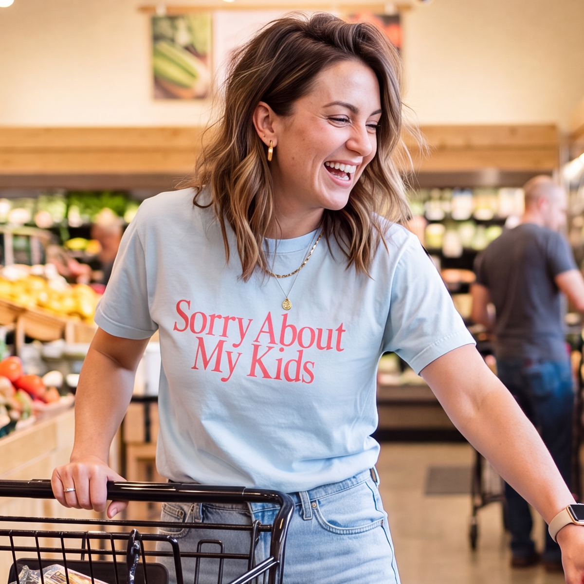 Lifestyle photo of a smiling woman pushing a shopping cart in a grocery store while wearing a light blue graphic T-shirt featuring the phrase "Sorry About My Kids" in large soft pink serif lettering across the chest. Styled with light wash jeans and simple gold jewelry, this funny parenting graphic tee is perfect for busy moms, errands, school pickup, grocery shopping, and everyday casual wear.