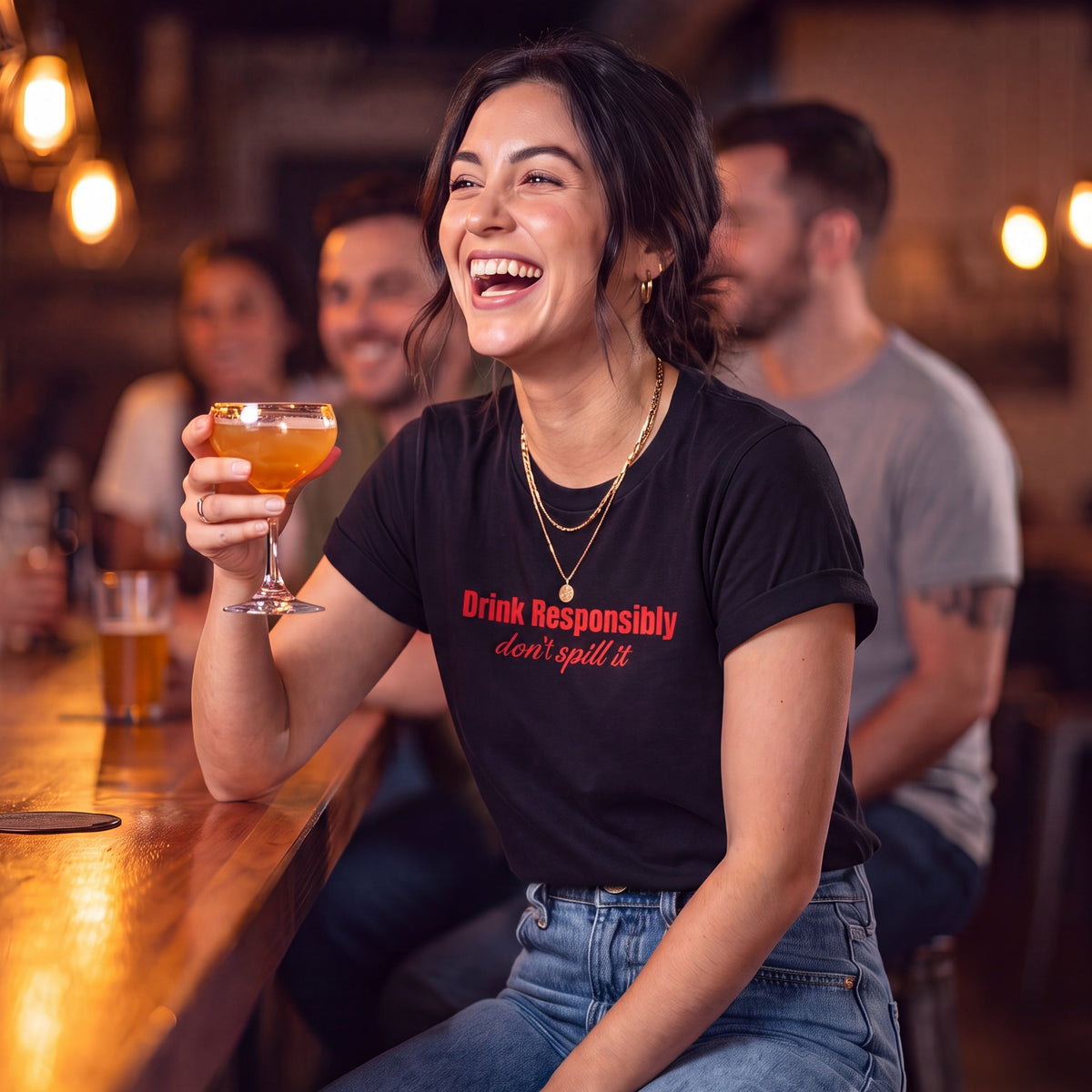 Smiling woman seated at a warmly lit bar wearing a black graphic t-shirt with red text reading “Drink Responsibly” and “don’t spill it,” while holding a cocktail. Friends and bar patrons appear softly blurred in the background, creating a casual nightlife lifestyle setting.