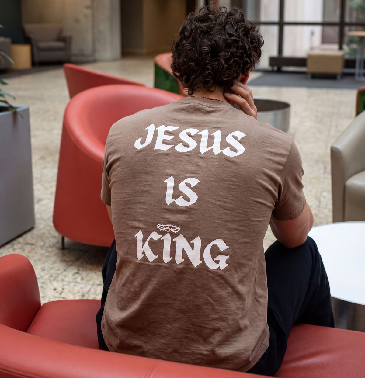 Man wearing a brown graphic t-shirt with large white back print reading “Jesus Is King,” seated in a modern indoor lounge.