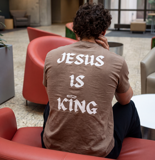 Man wearing a brown graphic t-shirt with large white back print reading “Jesus Is King,” seated in a modern indoor lounge.